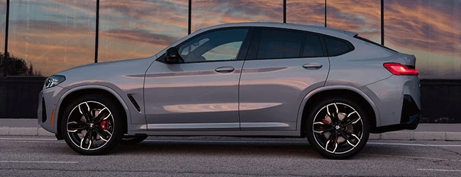 Side profile of a silver BMW X4 M40i parked at sunset with modern glass architecture reflecting the sky