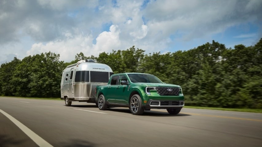 Green Ford Maverick towing a silver Airstream trailer on a highway surrounded by trees.