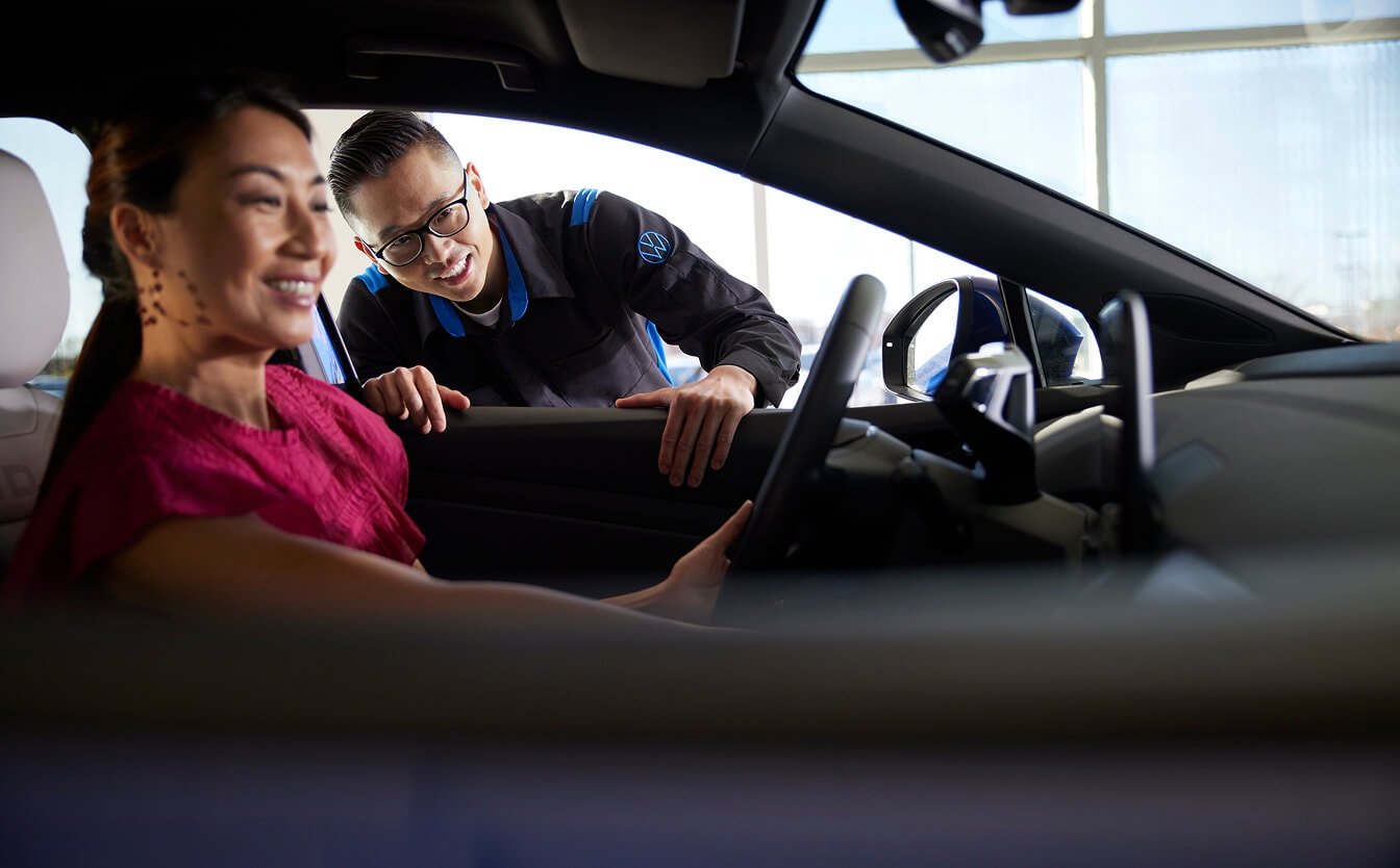 Volkswagen service advisor speaking with a customer seated inside a vehicle at a dealership, highlighting personalized service and assistance.