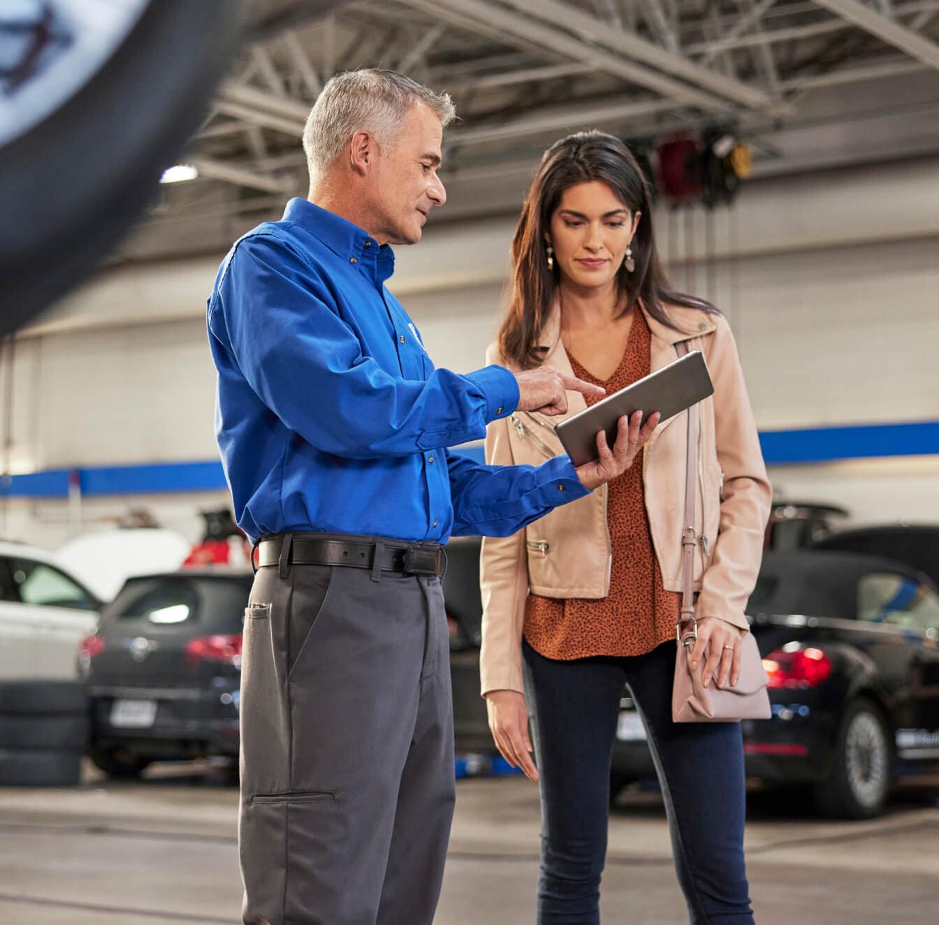 Volkswagen service advisor reviews coolant recommendations with a customer beside vehicles in a service bay.
