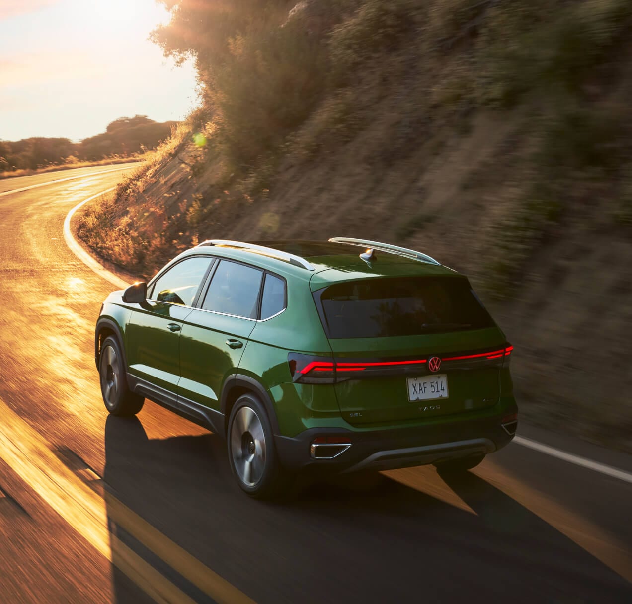 Rear three-quarter motion view of a green VW Taos SEL driving on a winding mountain road at sunset