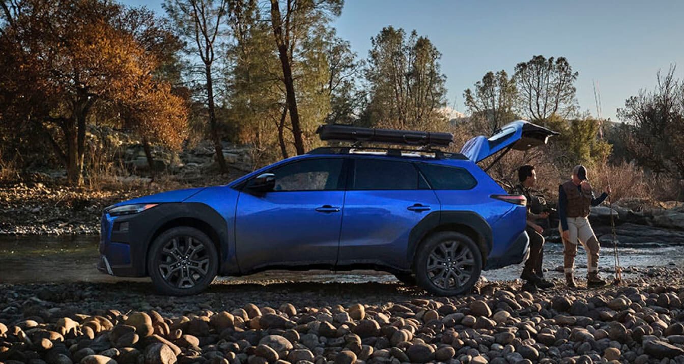 Side profile of a blue Subaru Trailseeker with an open hatch, parked on a rocky riverbank next to people with fishing pole.