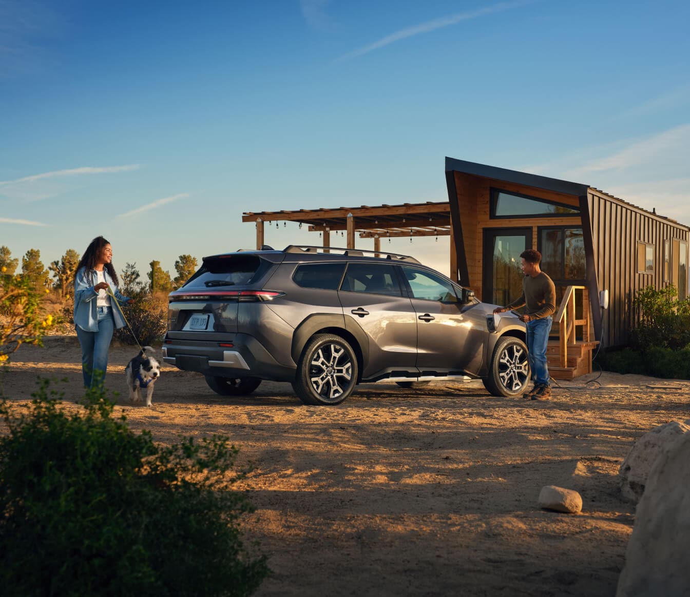 Side view of a grey Subaru Trailseeker charging outside a modern tiny home with people and a dog in a dirt yard.