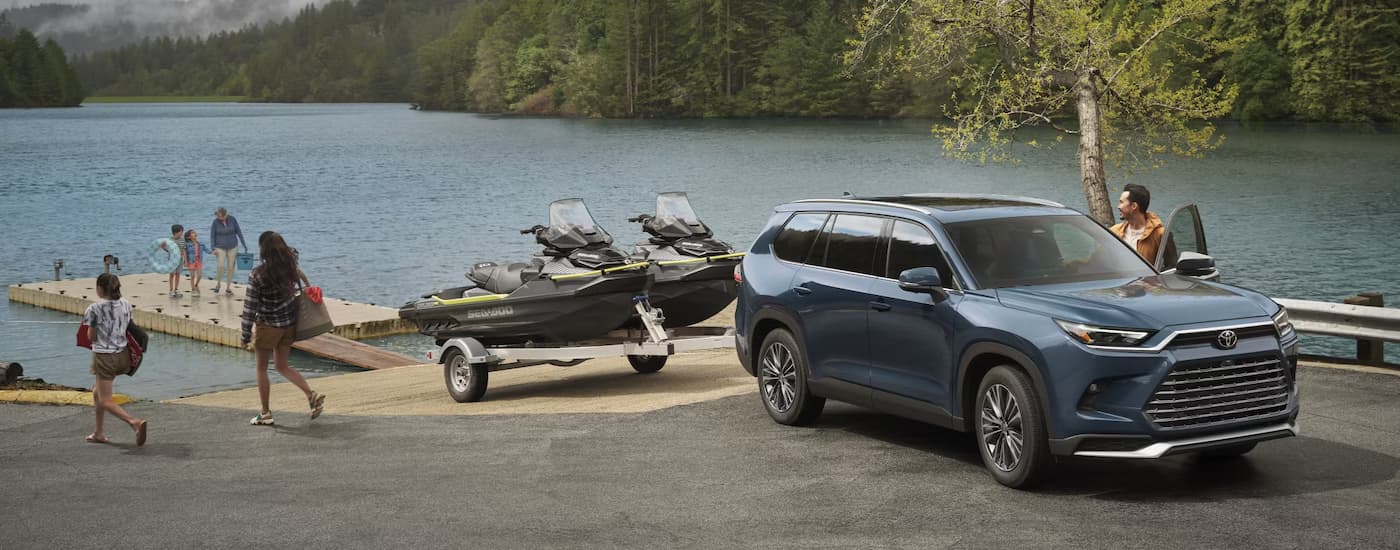 A blue 2025 Toyota Grand Highlander Platinum Hybrid Max is towing a trailer with two jetskis near a lake.