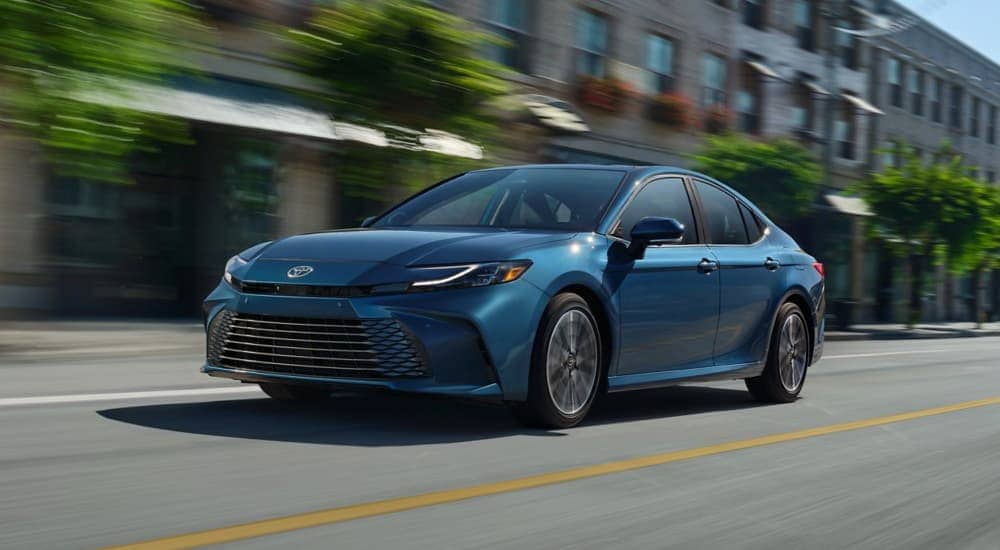 A blue 2025 Toyota Camry for sale is shown driving on a city street during a test drive.