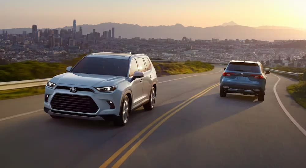A white and blue 2024 Toyota Grand Highlander are shown driving on a highway in opposite directions.