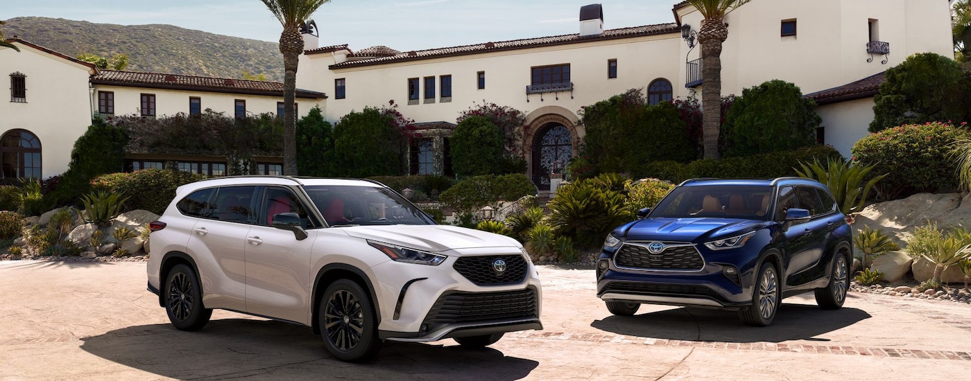 A white 2024 Toyota Highlander XSE and a blue 2024 Highlander Hybrid Platinum parked outside a villa.
