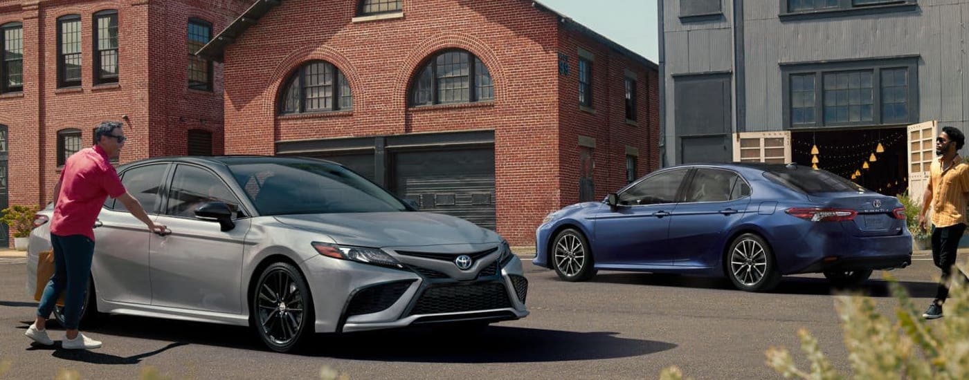 A blue and a silver hybrid 2024 Toyota Camry XSE for sale are parked outside of a brick building.