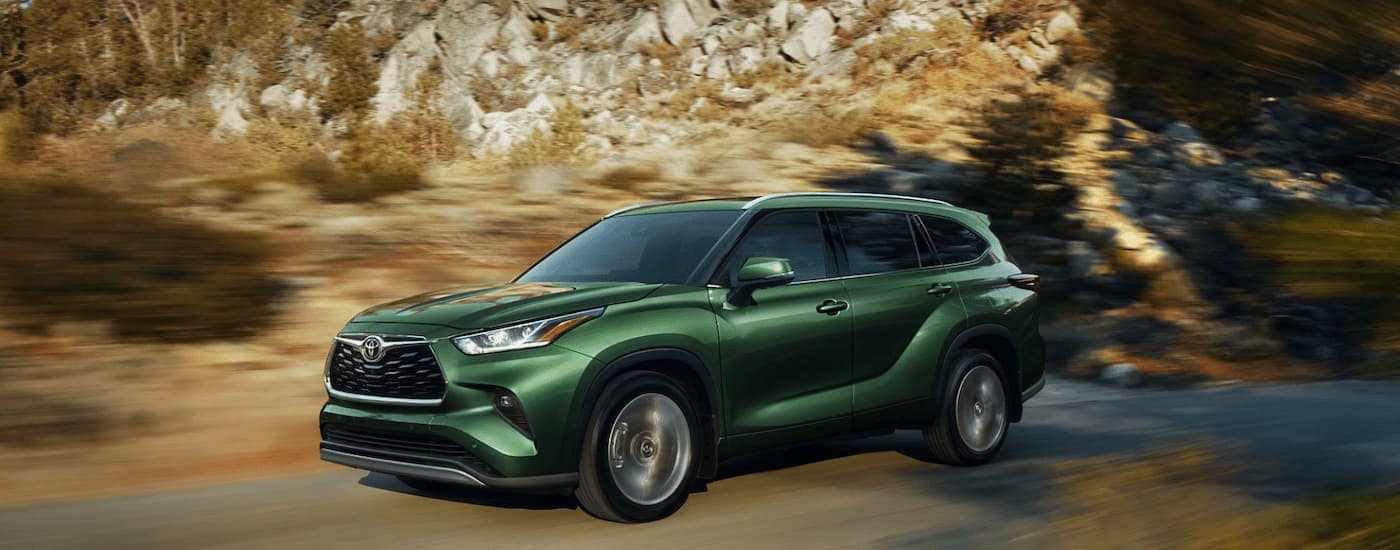 A green 2023 Toyota Highlander is shown driving on a road next to a rocky area.