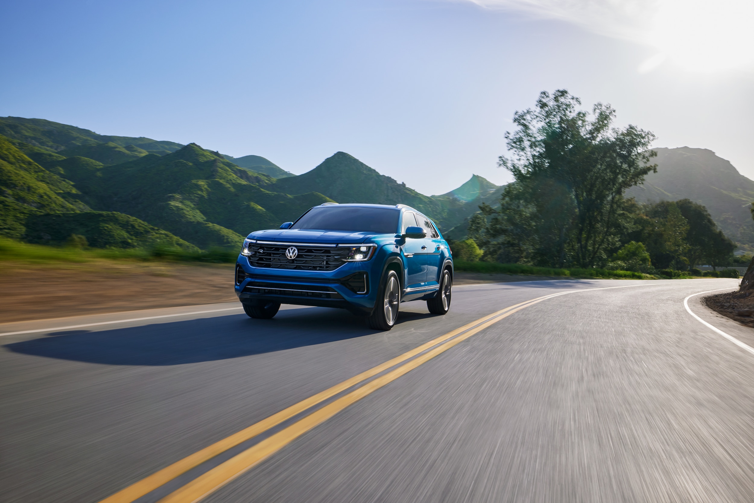 blue Volkswagen Atlas Cross Sport driving past green hills in the background