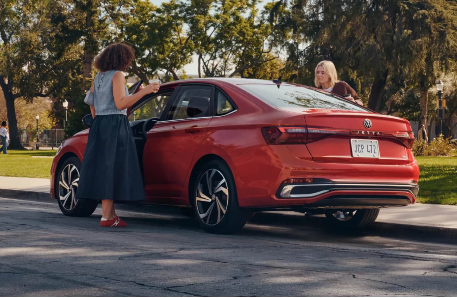 Rear three-quarter view of two women near a 2027 Volkswagen Jetta SEL in Kings Red Metallic.