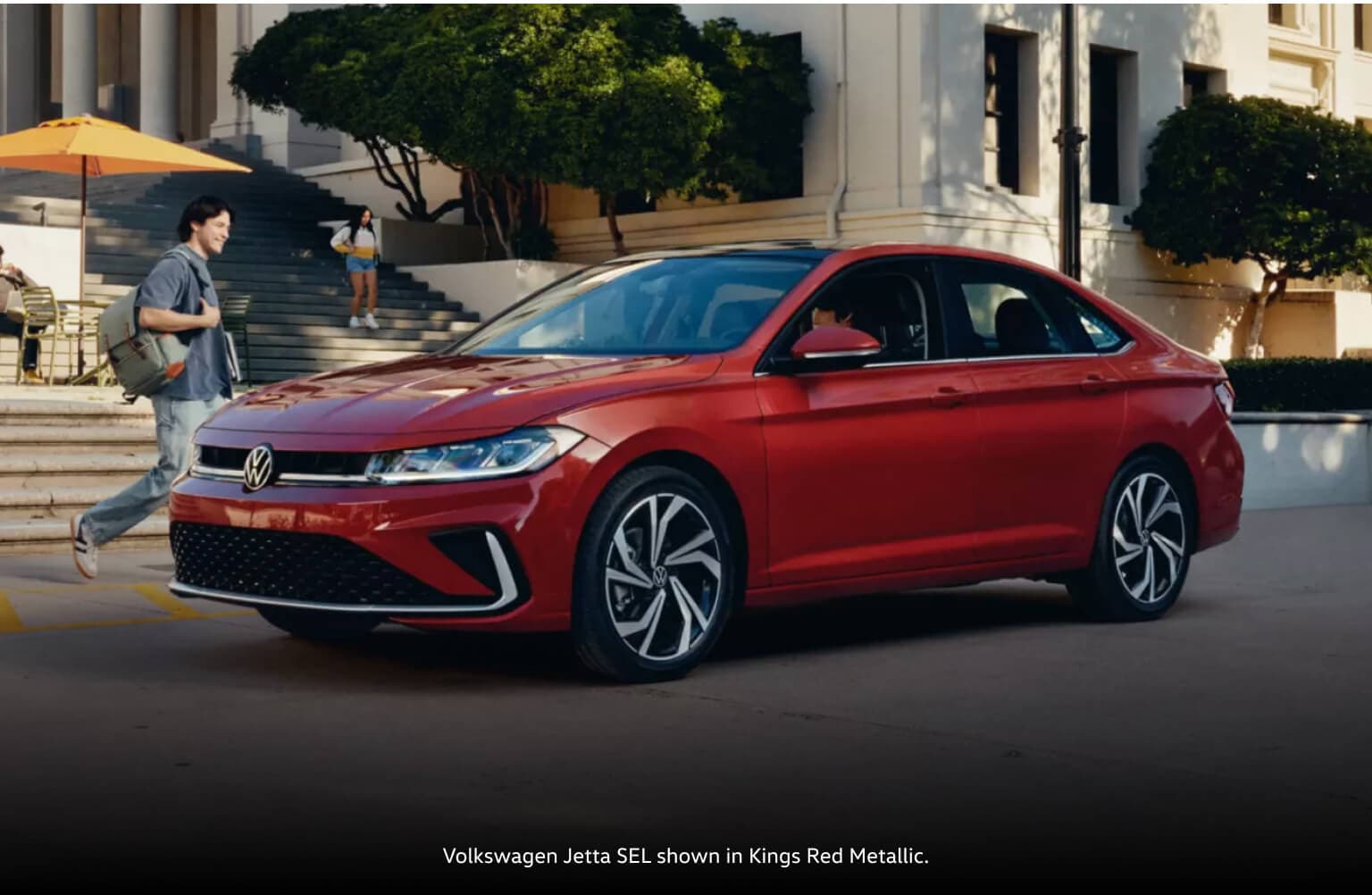 Front three-quarter view of a 2027 Volkswagen Jetta SEL in Kings Red Metallic near students.