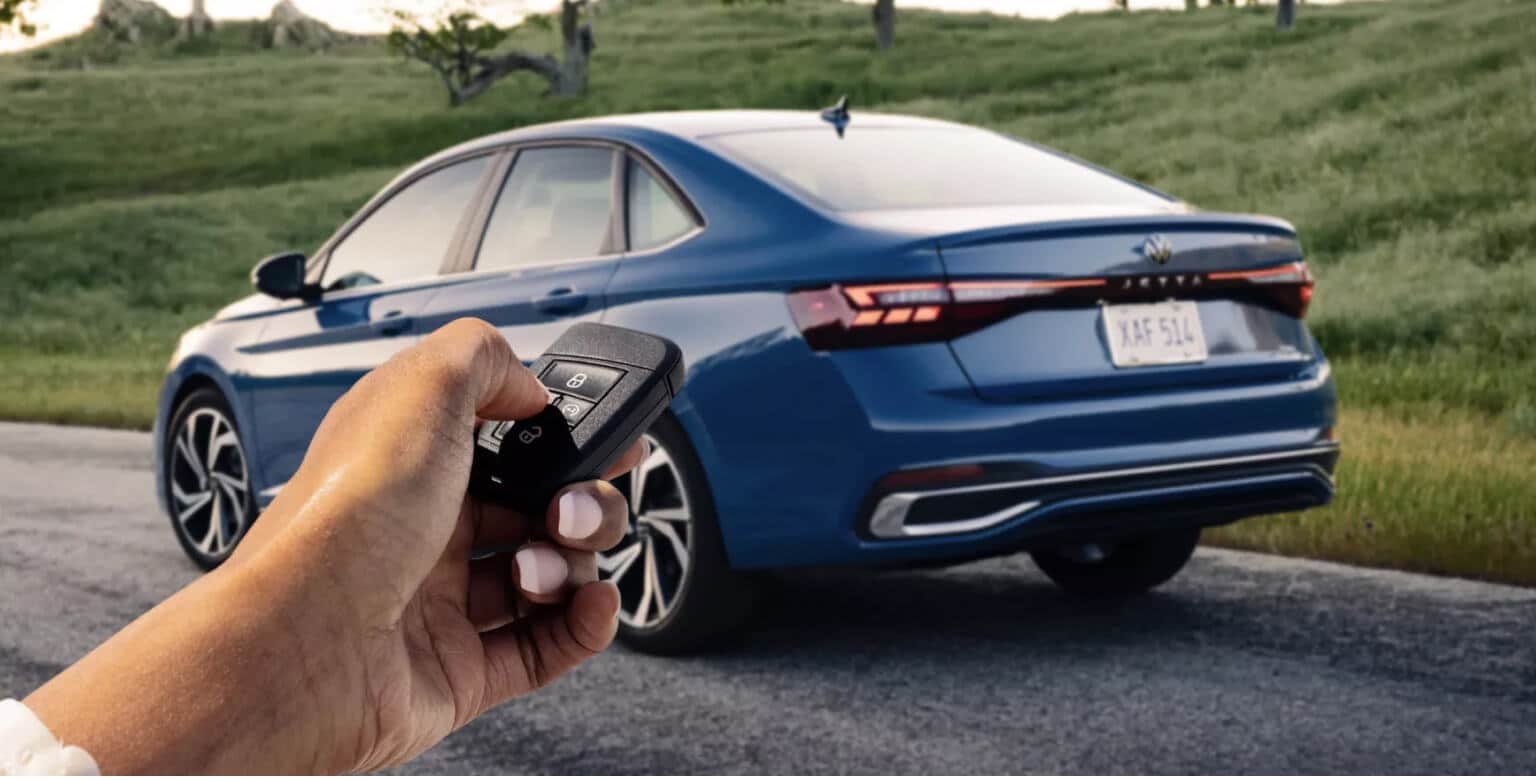 Close-up of a hand holding a key fob aiming at a 2027 Volkswagen Jetta SEL in Monterey Blue Pearl.
