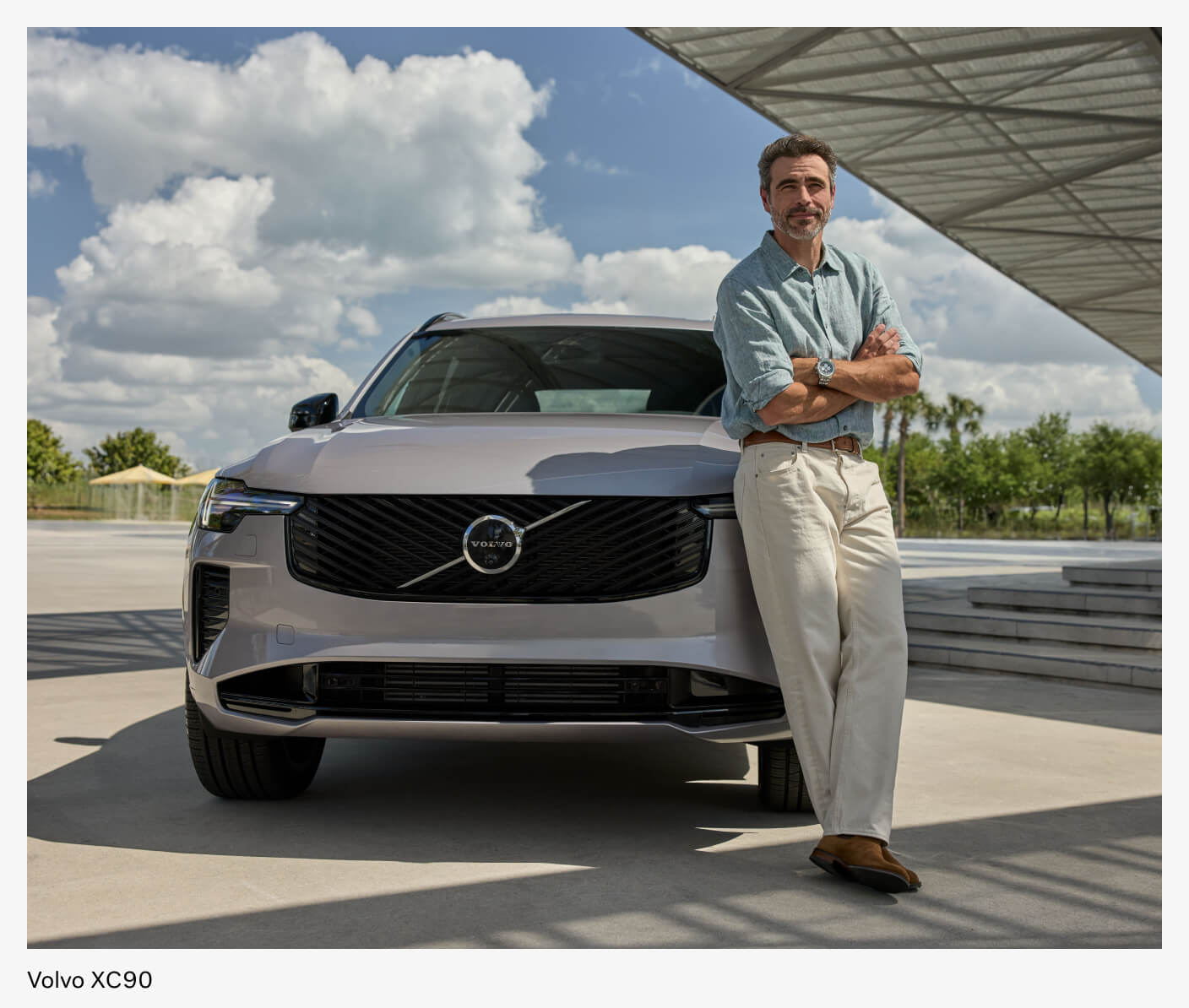 A man confidently leans on his parked Volvo XC90 in a modern waterfront lot.