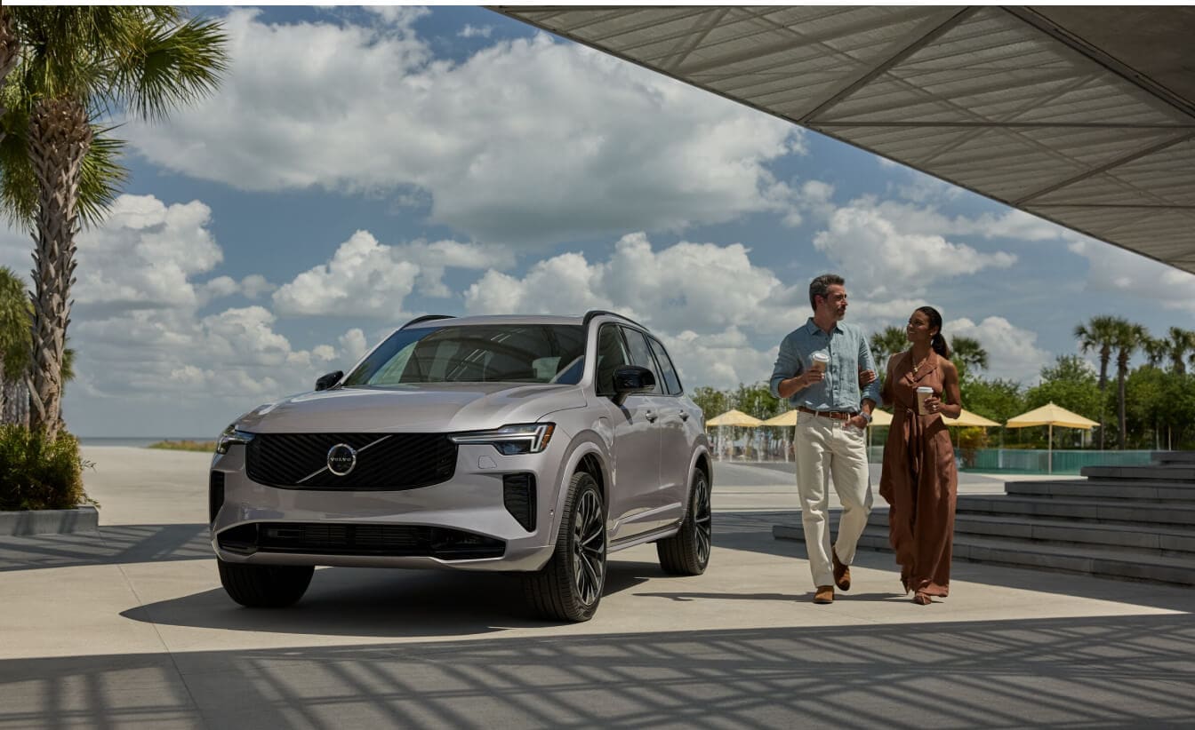 Front three-quarter view of a silver Volvo XC90 SUV parked in a plaza with a walking couple.