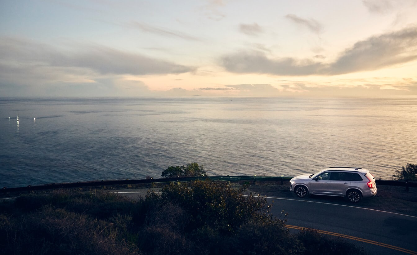 High-angle side view of a silver Volvo XC90 SUV driving on a coastal road overlooking the ocean.