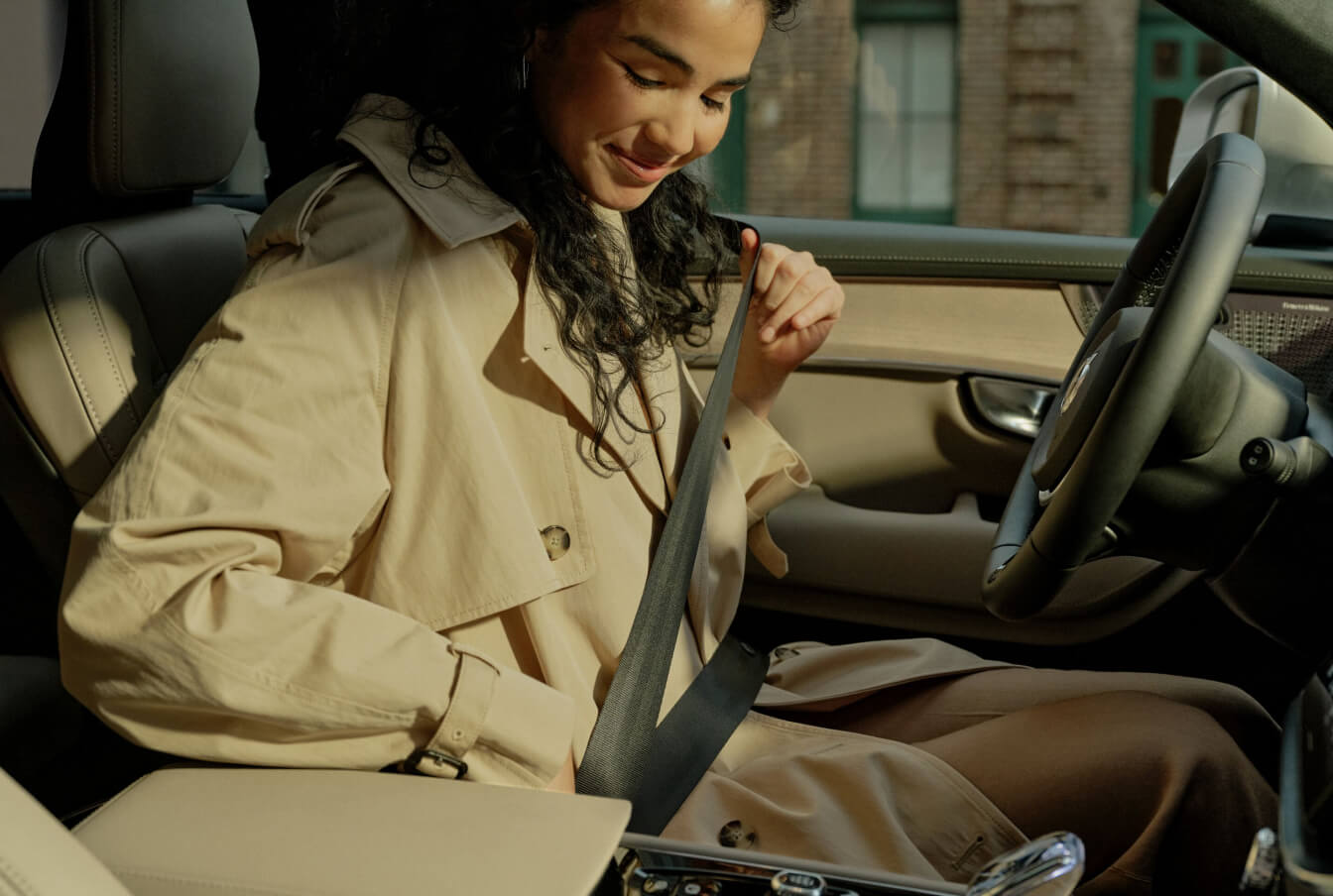 Interior view of a driver fastening her seatbelt inside a tan Volvo XC90 cabin.