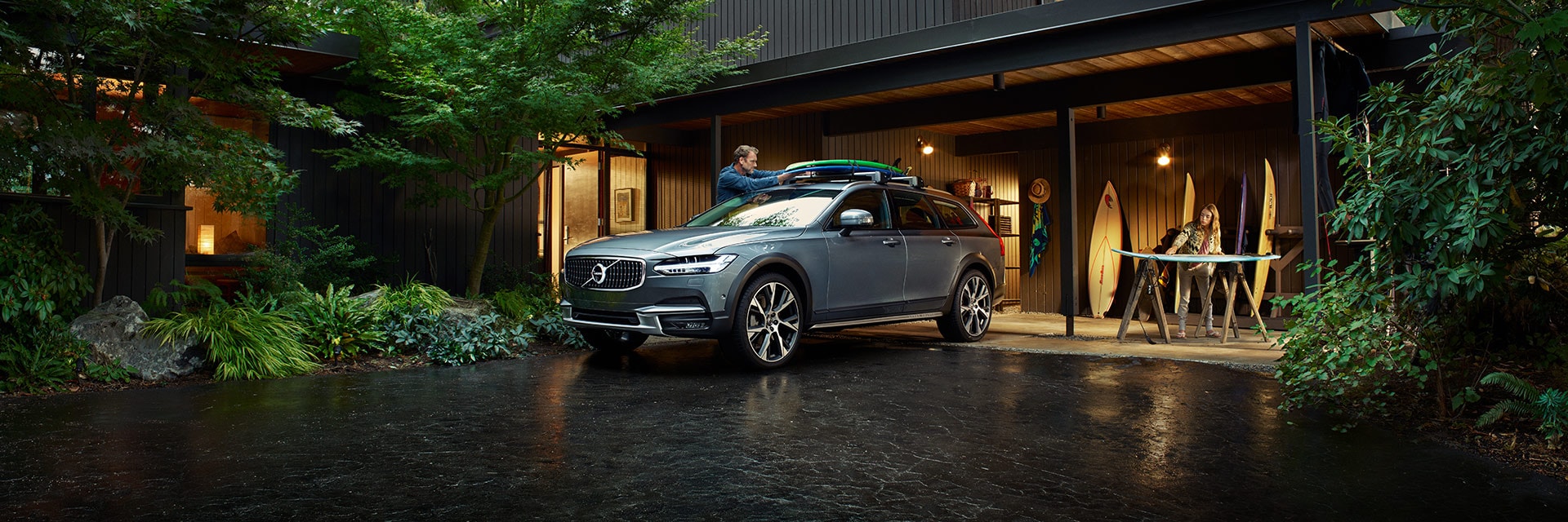 Volvo Cars New Bern is a Volvo Dealership in New Bern near Jacksonville NC | Man tying surfboards to the roof of a Silver MY20 Volvo V90 on a rainy day in his driveway