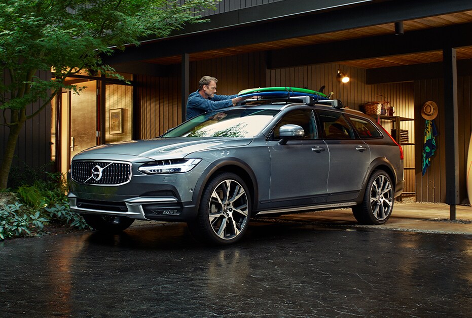 Volvo of New Bern is a Car Dealership in New Bern near Hubert NC | Man packing kayaks onto Gray 2020 Volvo V90 in front of house in the woods