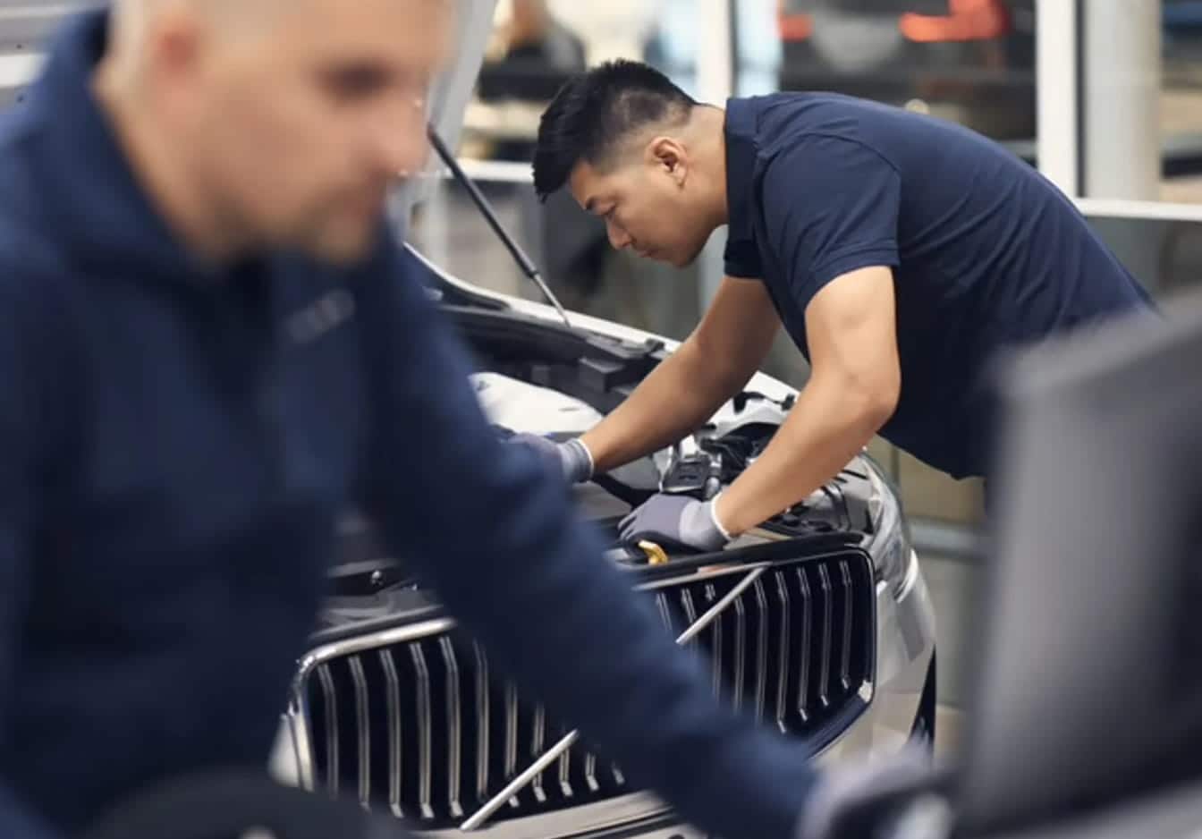 Volvo technician inspecting the engine bay of a Volvo SUV inside a service center with another technician blurred in the foreground.