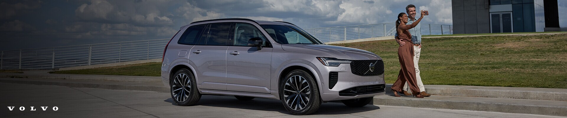 Three-quarter front view of a luxury Volvo XC90 Ultra in Bright Dusk Metallic as a couple walks by on a sunny paved boardwalk.