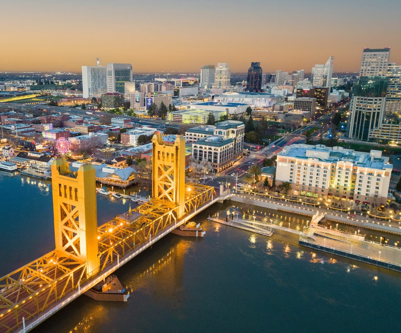 Aerial view of a city skyline at sunset featuring a river, a lit suspension bridge, and waterfront buildings.