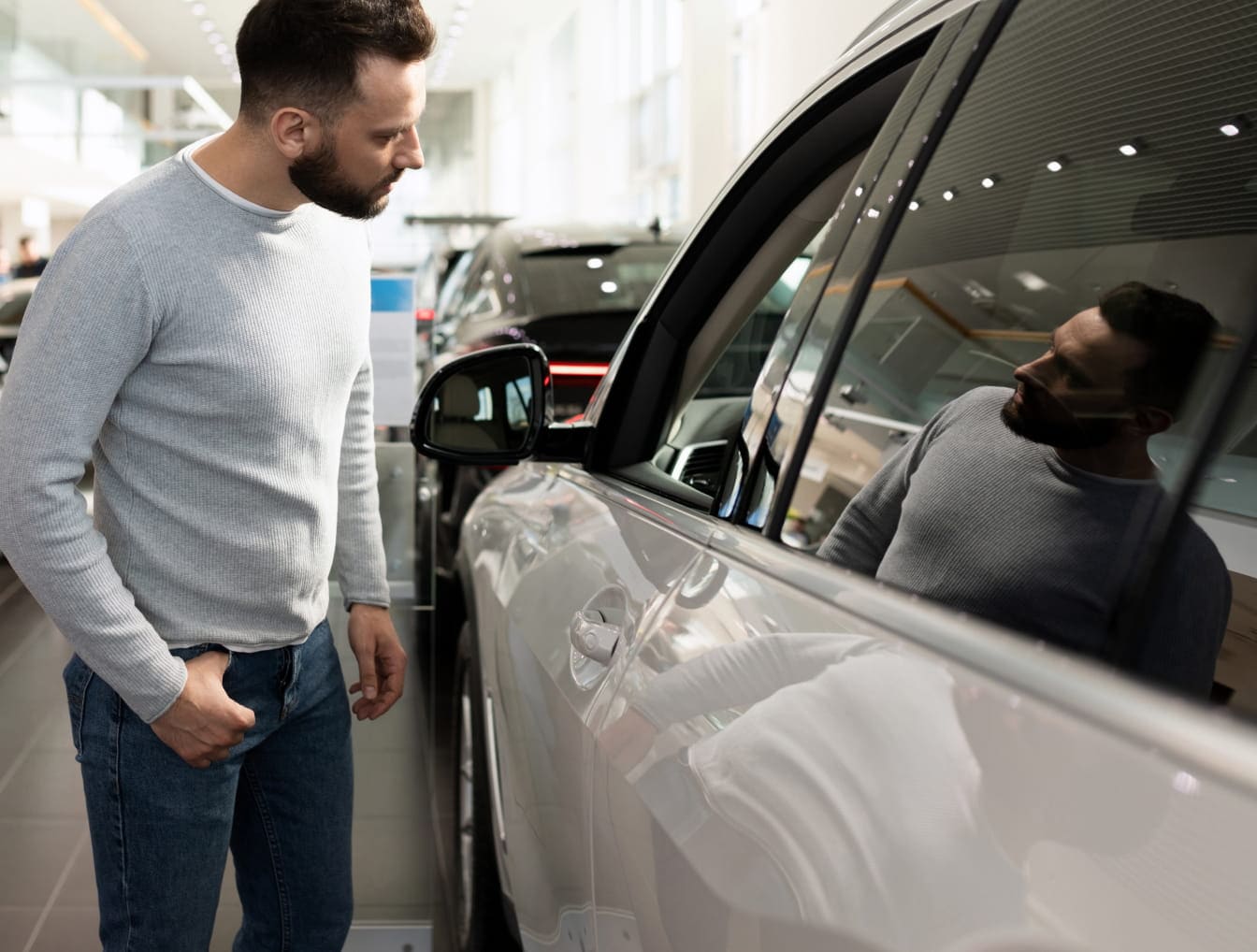 Man inspecting the side of a white vehicle SUV inside a bright dealership showroom with other vehicles in the background.