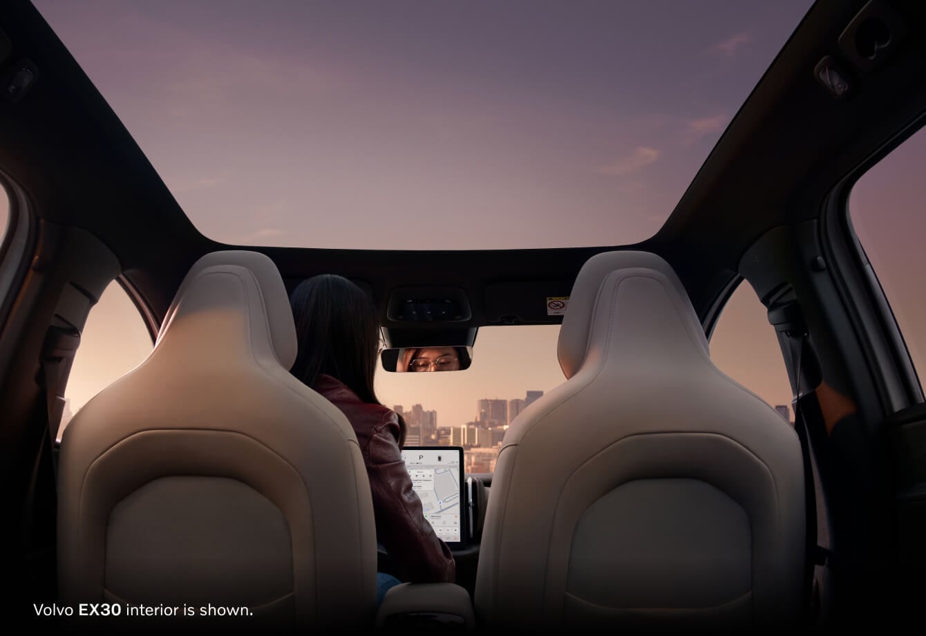 Volvo EX30 interior is shown from the back seats with female driver viewing the infotainment and city in background.