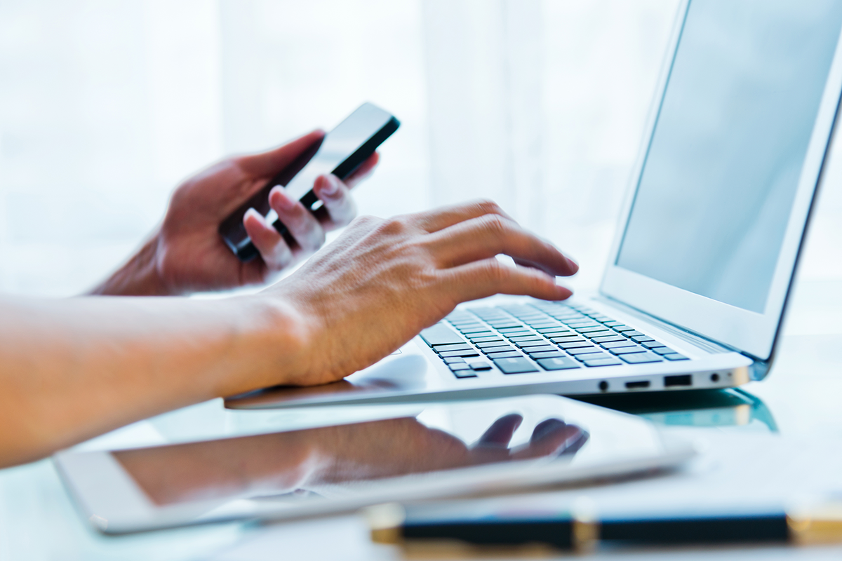 A woman typing on a laptop while also holding her phone