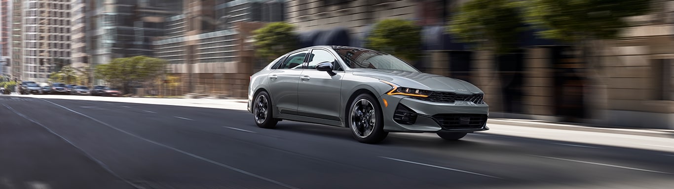 Best MPG At   West Broad Kia in Henrico, VA | Grey 2023 Kia K5 driving down city road during the day 