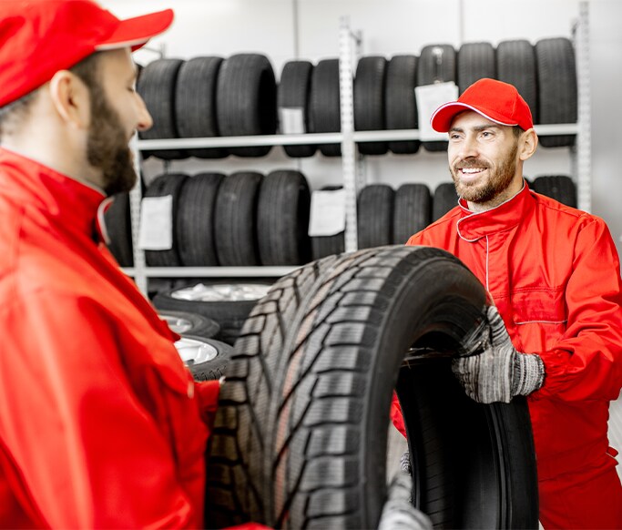 How to Get Your Car Summer Ready at West Broad Kia in Henrico, VA | One mechanic passing a new tire over to another mechanic, both in red uniforms
