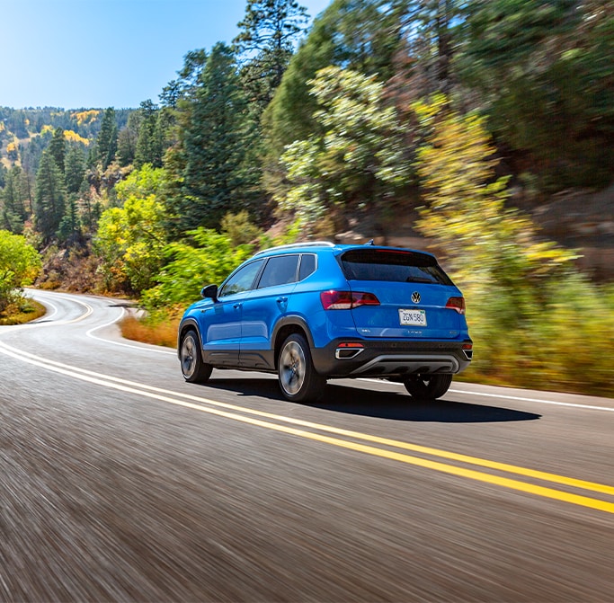 Best MPG At West Broad VW in Richmond, VA | Rear view of a blue 2023 Volkswagen Taos driving down the road with tall trees in the background 