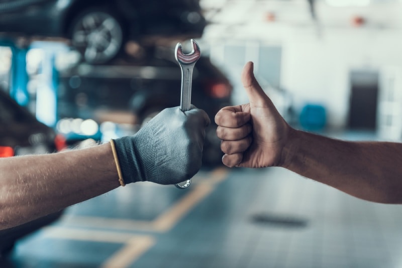 Mechanic holds a wrench perpendicular to his arm in a gloved hand while person having her car serviced puts her hand into the thumbs up position next to the mechanic’s hand