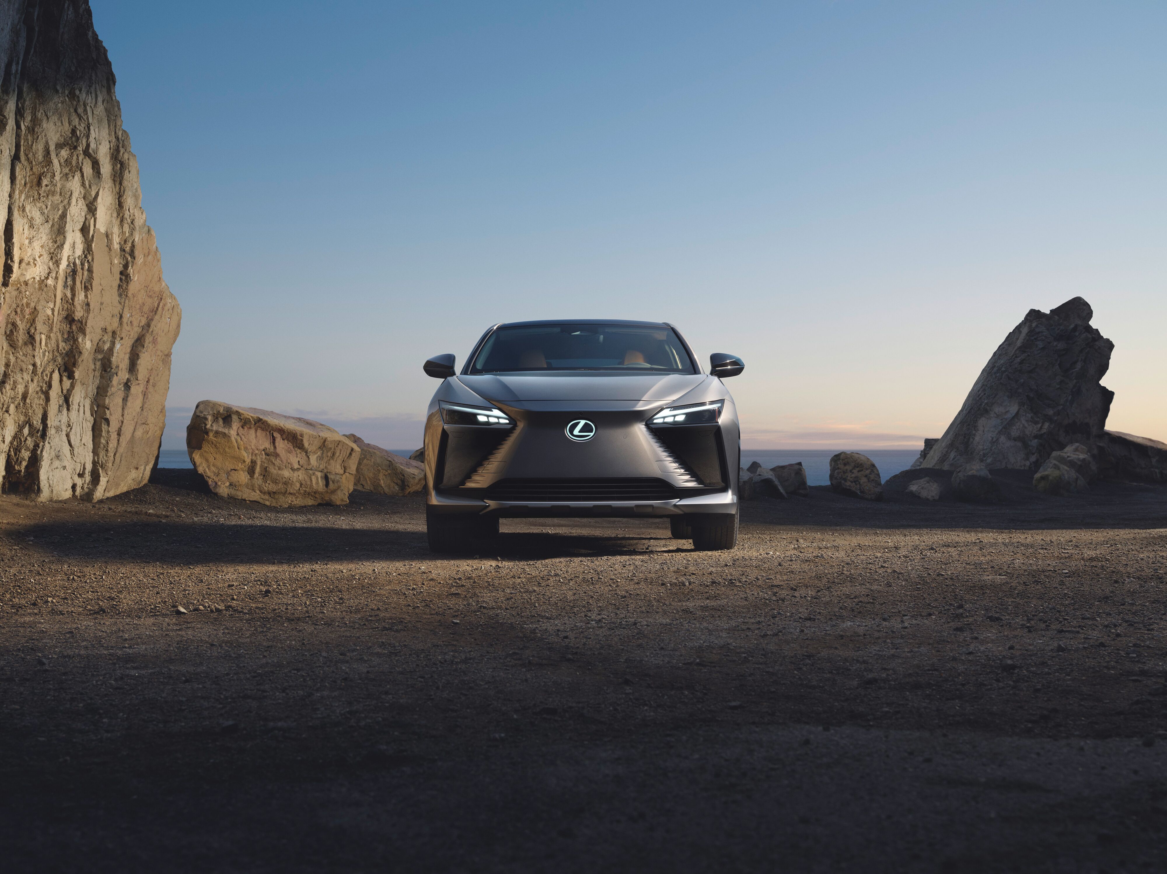 Direct front view of a gray Lexus RZ electric SUV parked on gravel between large rocky formations at dusk.