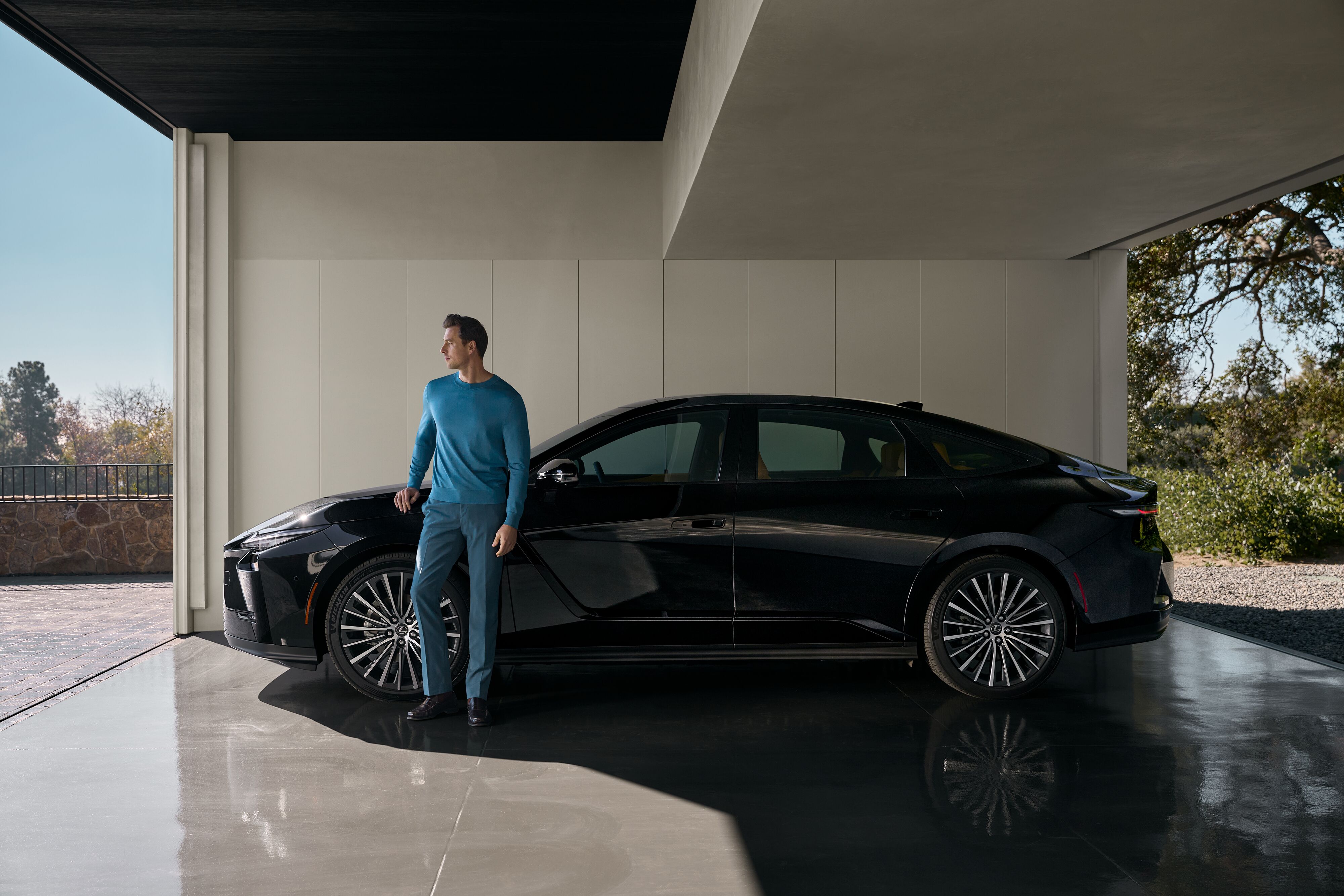 Side profile view of a black Lexus electric luxury sedan with an elegantly dressed man standing by the car under a modern pavilion.