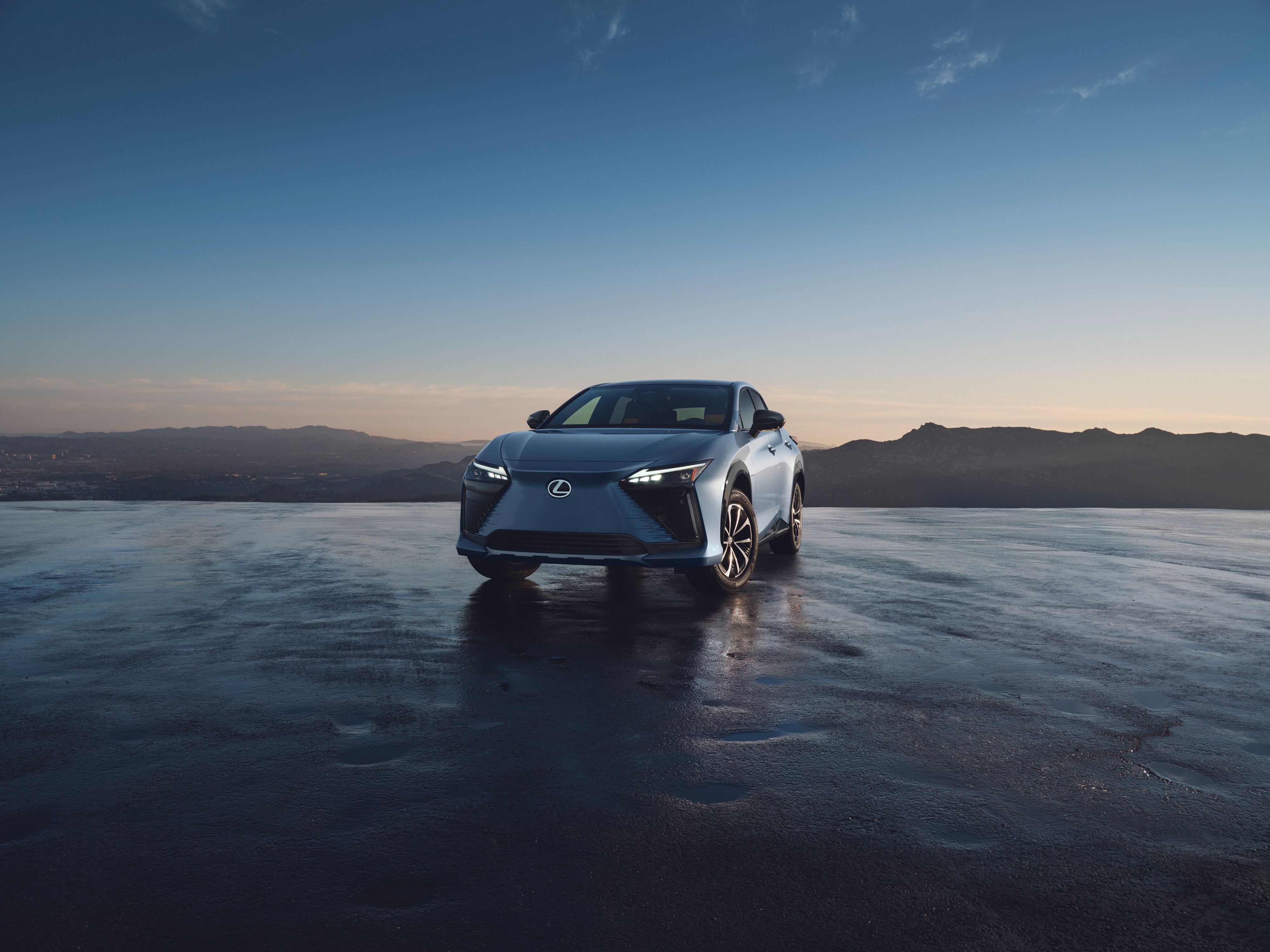 Lexus RZ electric SUV parked on wet pavement with scenic mountain landscape at sunrise.