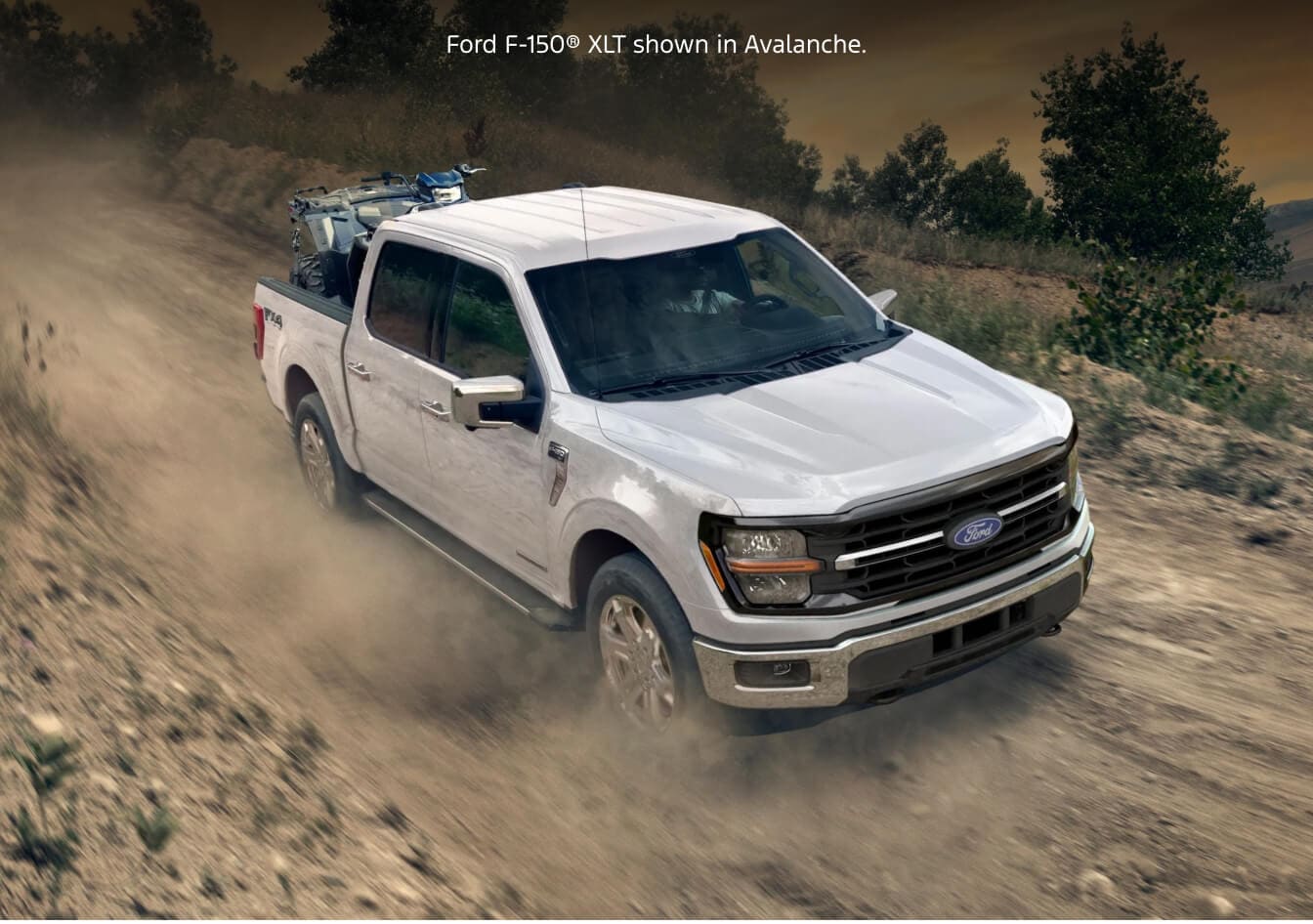 High-angle dynamic action shot of a 2026 Ford F-150 XLT in Avalanche hauling an ATV up a dusty dirt trail.