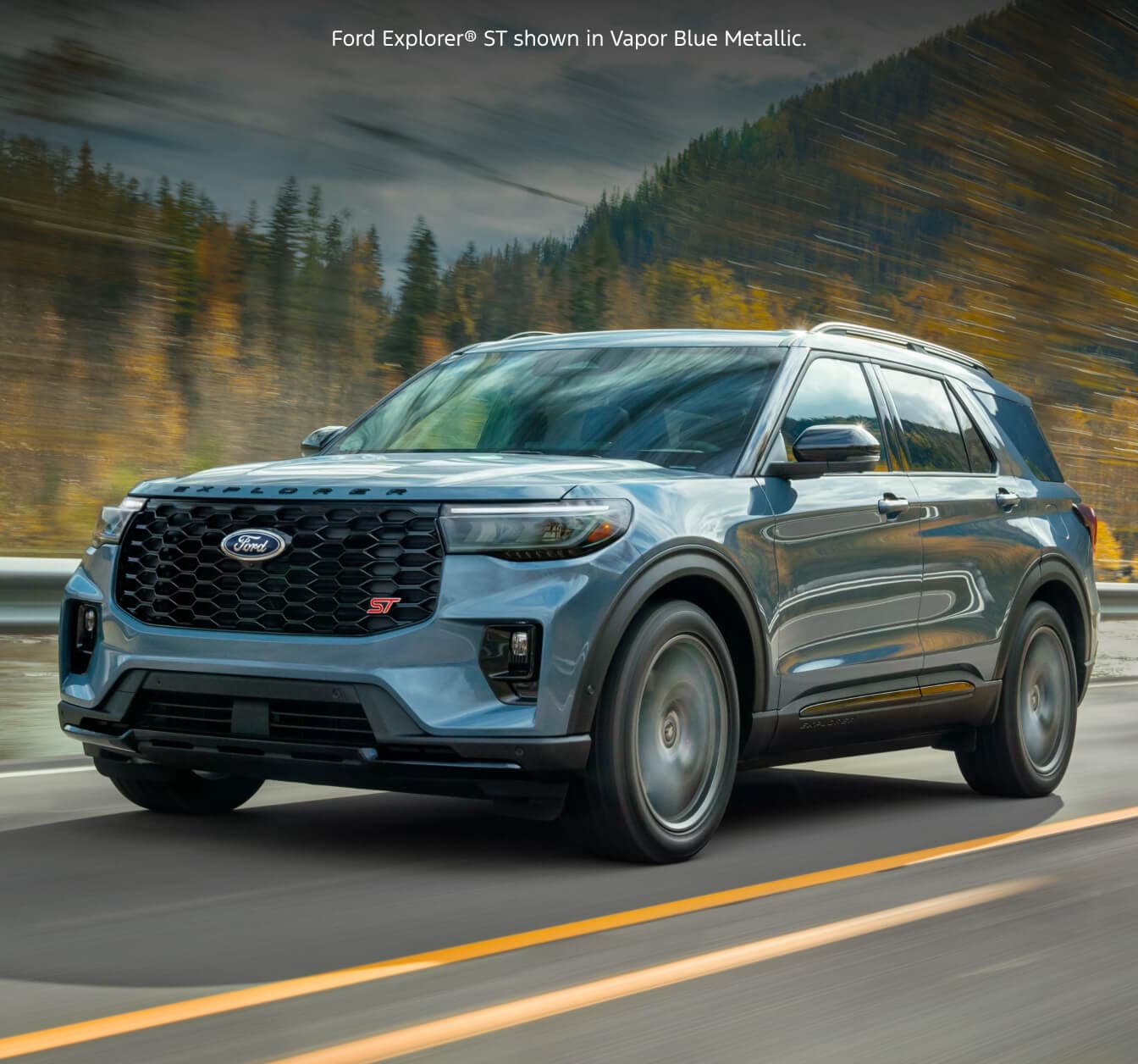 Front three-quarter shot of a 2026 Ford Explorer ST in Vapor Blue Metallic driving along a highway against a forested mountain.