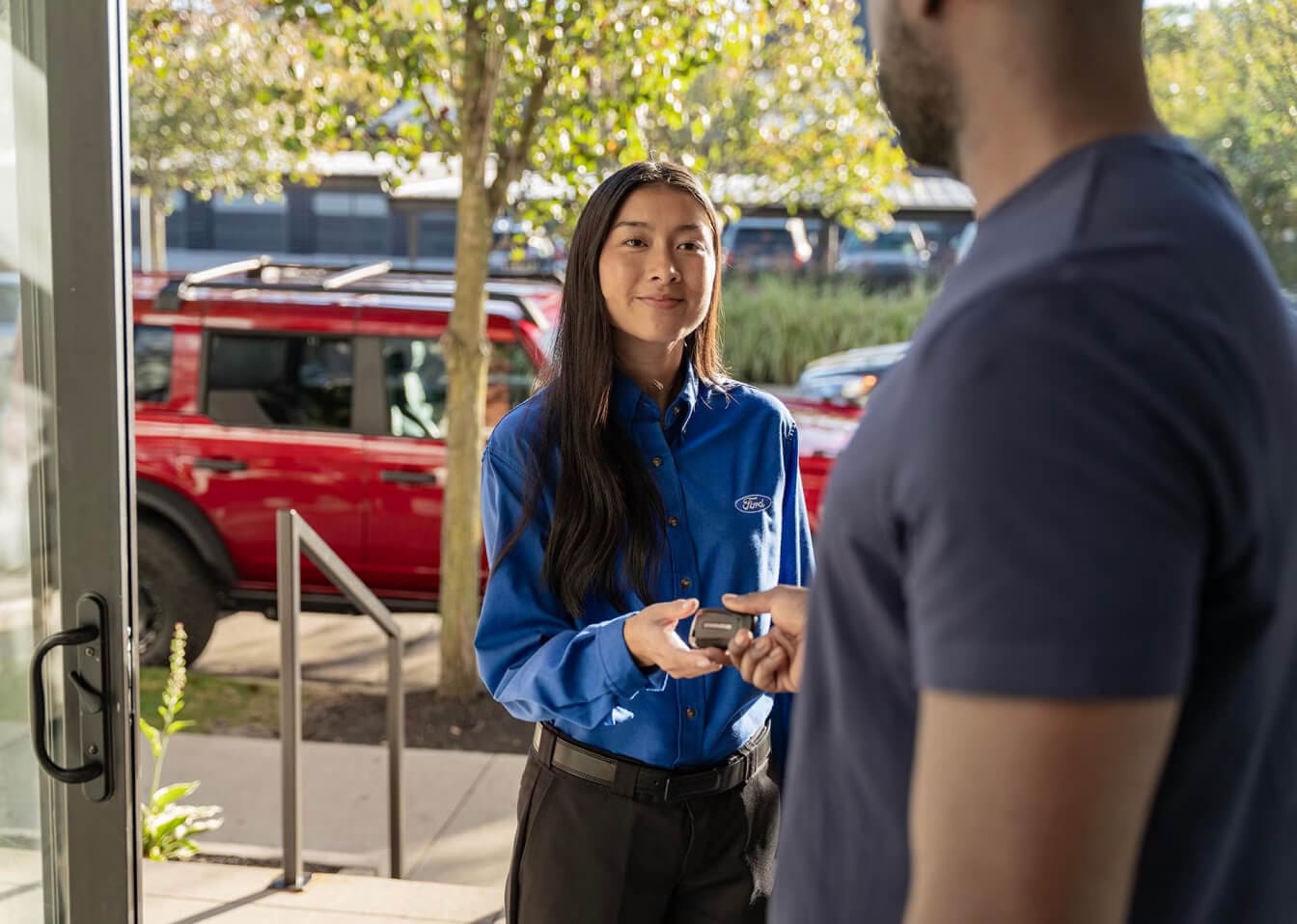 A Ford Representative handing the key fob to a customer at their doorstep, with a 2026 Ford Bronco Outer Banks in Hot Pepper Red Metallic.
