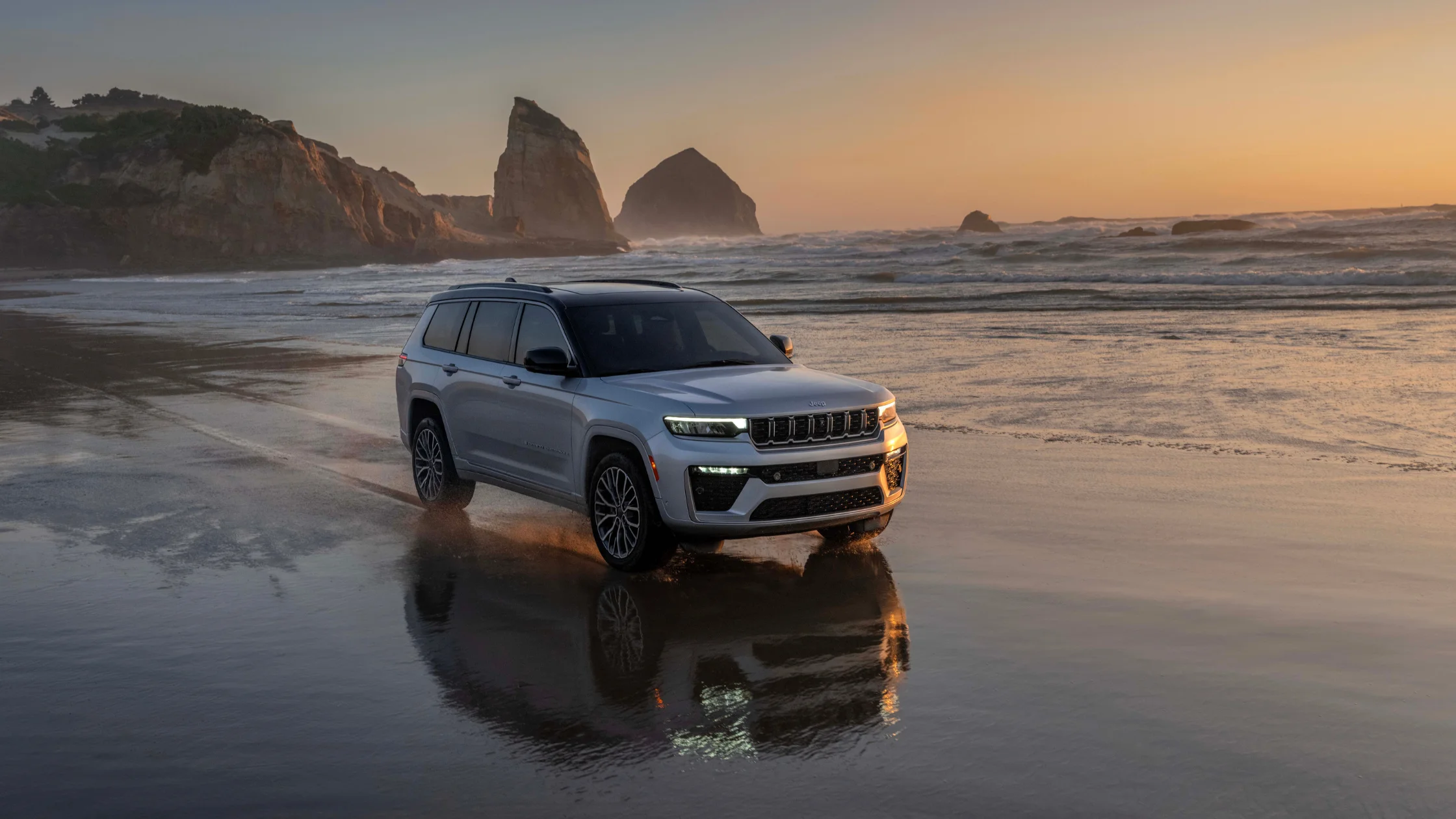 2026 Jeep® Grand Cherokee L driving on wet beach at sunset with ocean rocks and LED lights