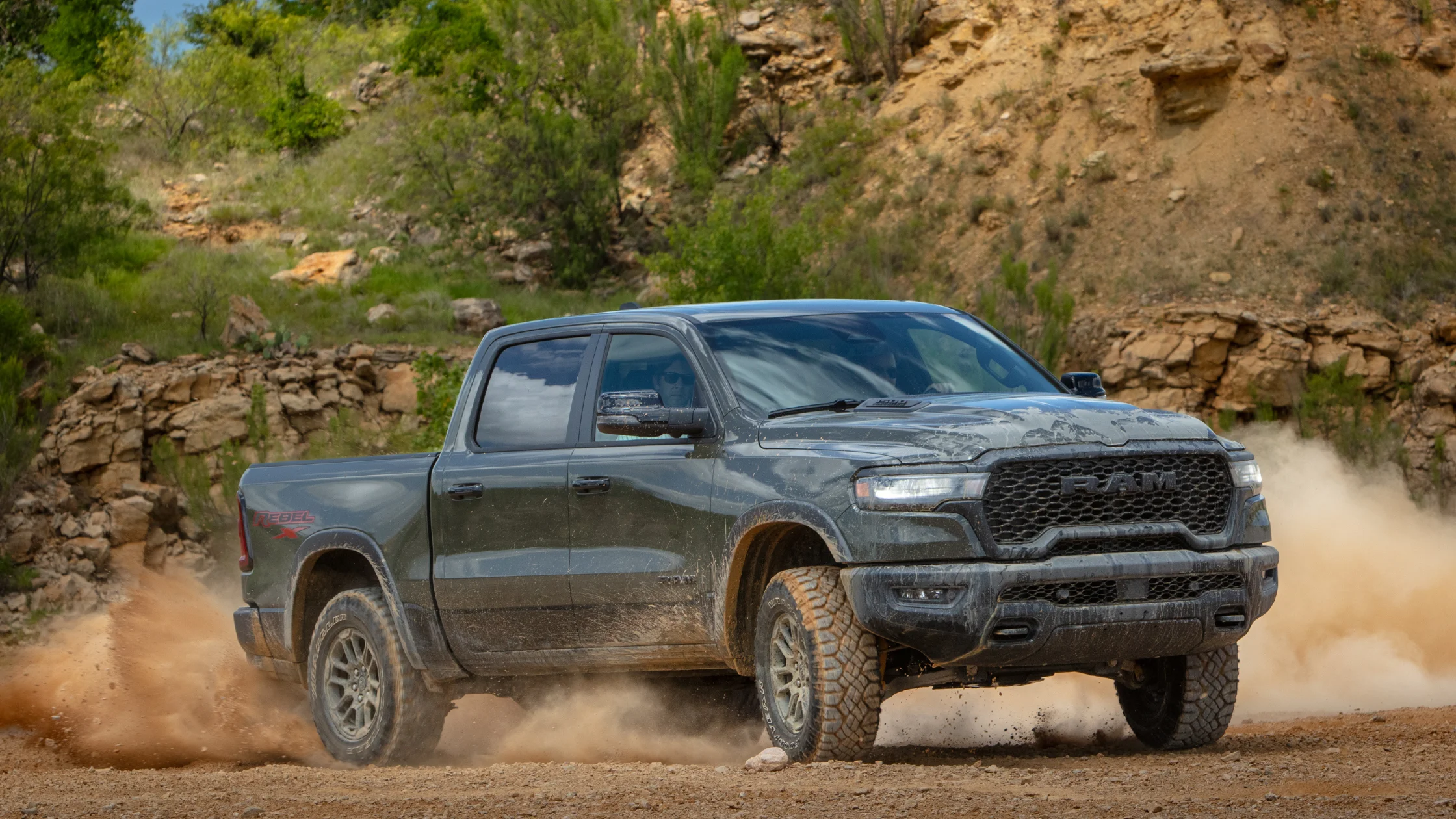 2026 Ram 1500 Rebel gray pickup powering through dusty off-road trail with rugged tires and rocks