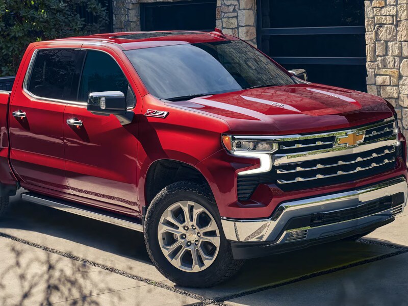 A shiny red pickup truck with a bold grille and chrome accents is parked on a driveway in front of a stone building, exuding power and elegance.