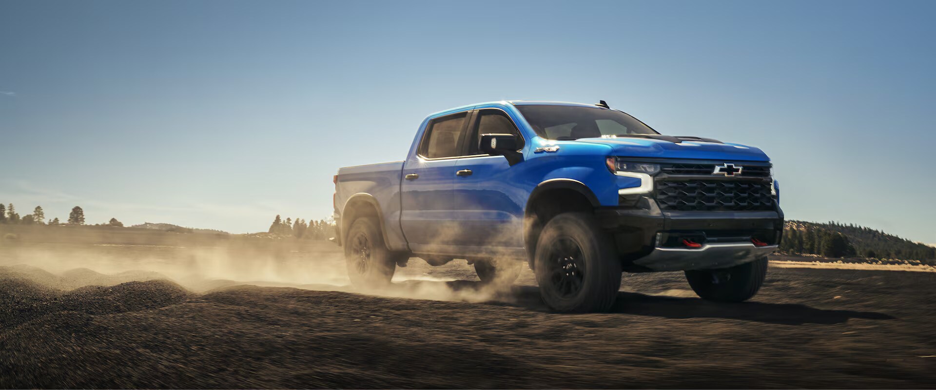 Blue Chevrolet Silverado pickup truck driving off-road on dusty terrain under clear sky.