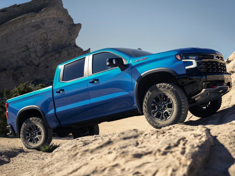 Blue Chevrolet ZR2 pickup truck climbing rocky terrain with large boulders and clear sky background.