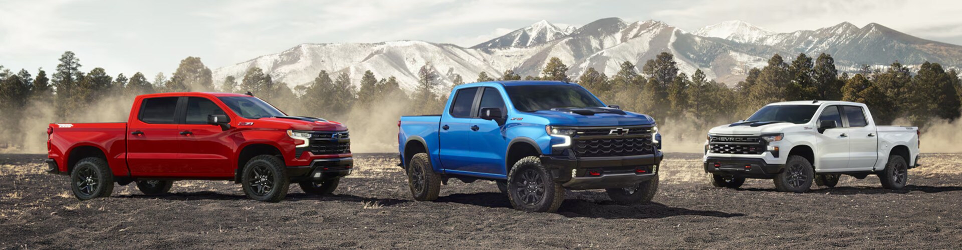 Three pickup trucks—red, blue, and white—are parked on rugged terrain with snow-capped mountains and pine trees in the background, conveying a sense of adventure.