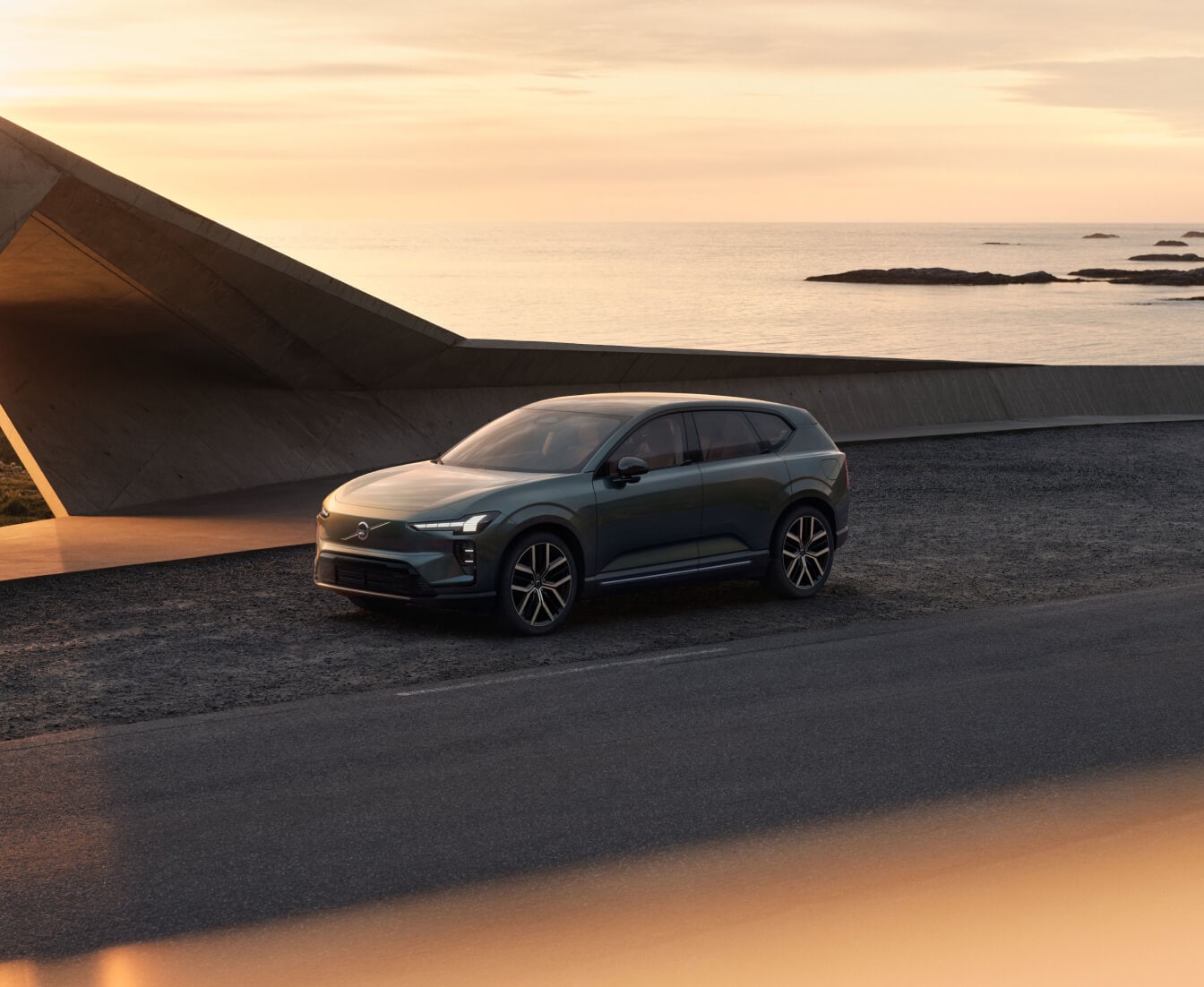 Front three-quarter view of a green Volvo EX60 parked on a coastal road by modern concrete architecture at sunset.