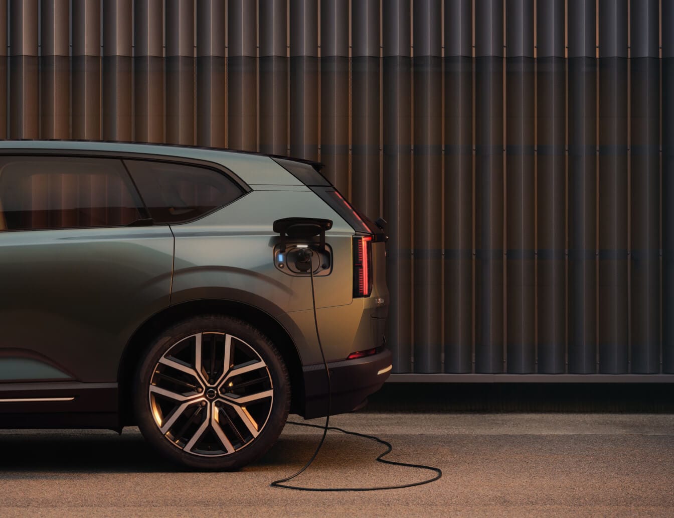 Rear side view of a green Volvo EX60 charging, parked in front of a corrugated wall at sunset.