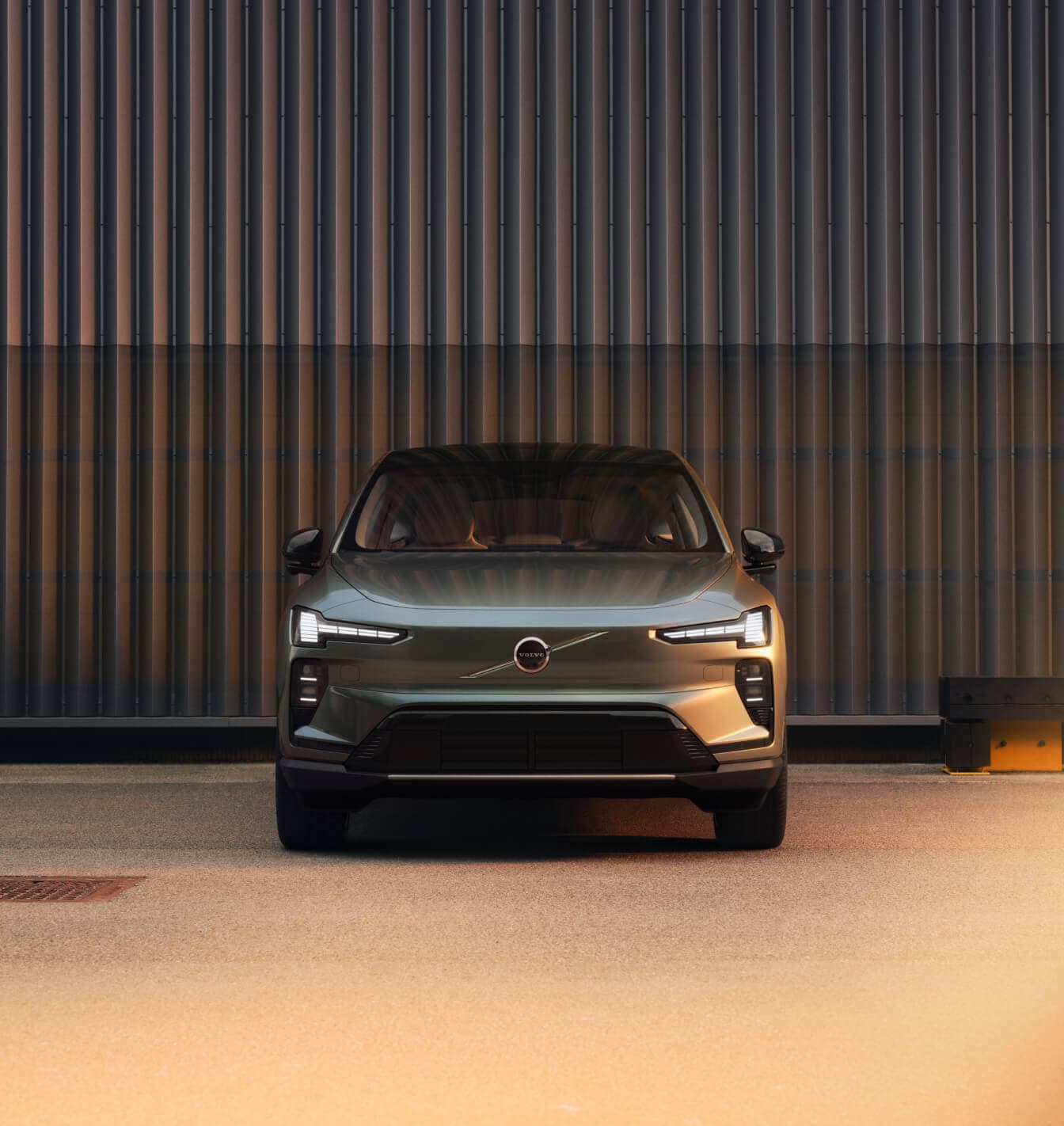 Front view of a green Volvo EX60 parked in front of a corrugated wall at sunset.